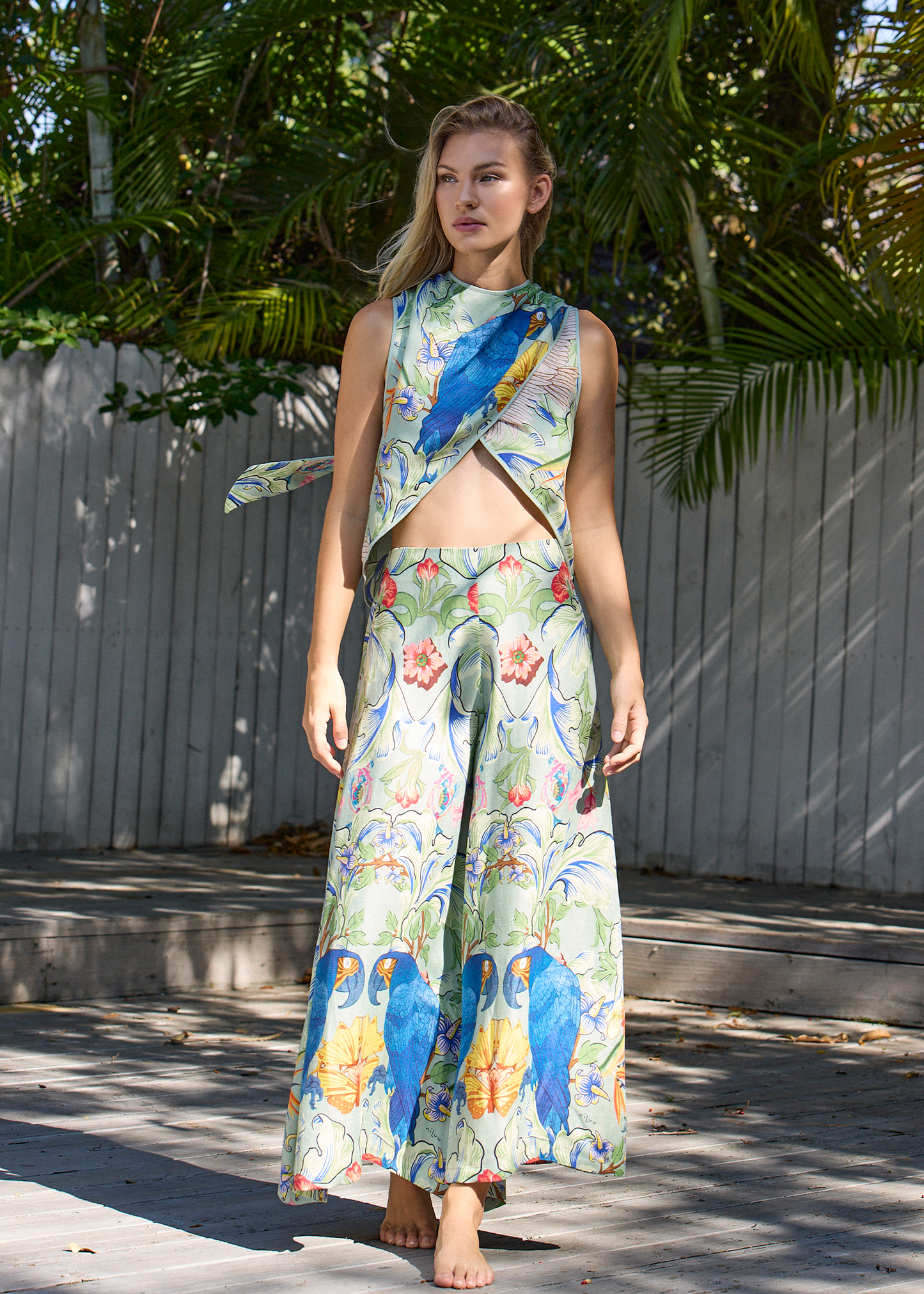 Front view of the tropical printed co-ord set worn outdoors. Sleeveless wrap-style top with midriff cutout and matching wide-leg pants featuring bold parrot and floral motifs.