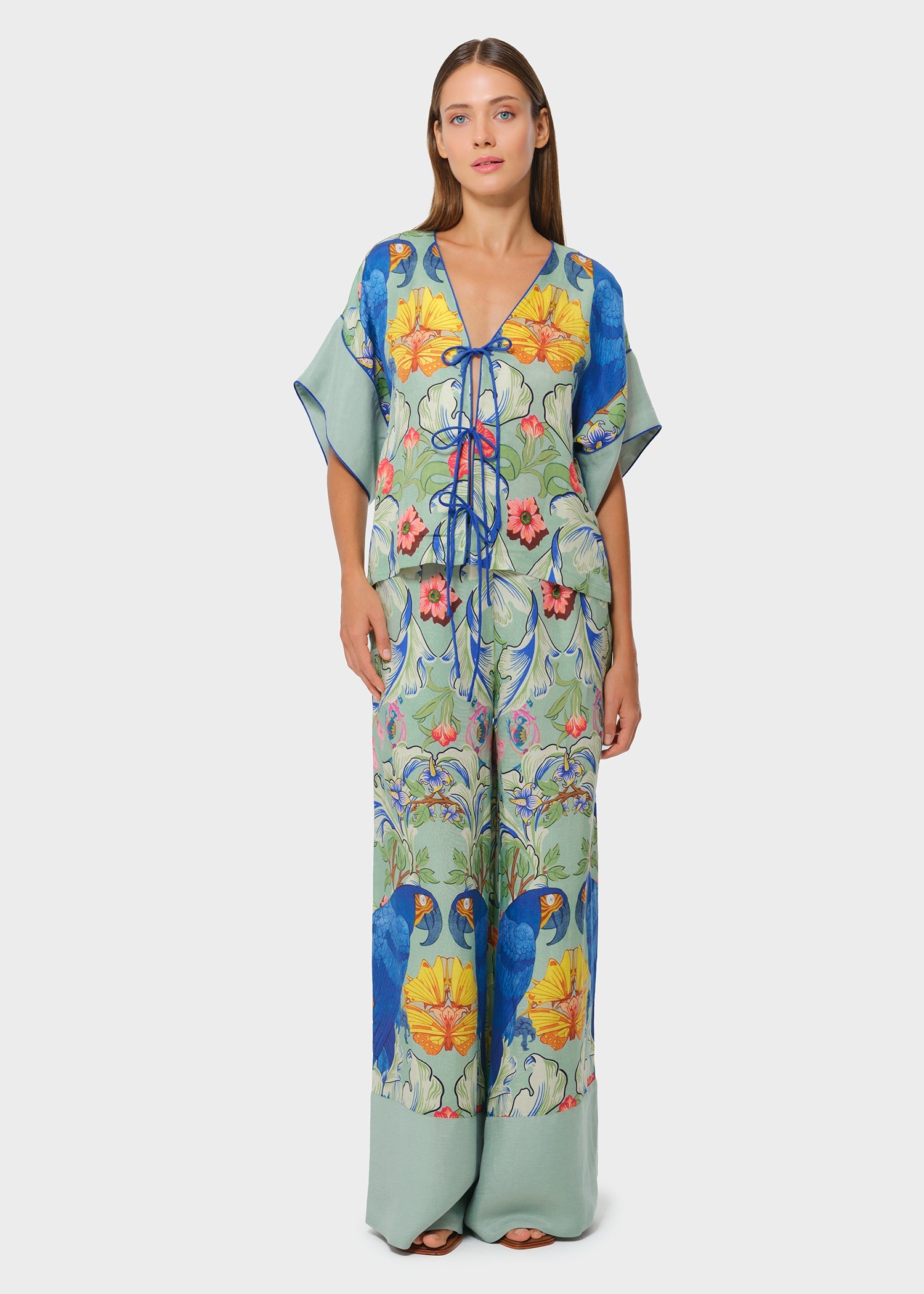 Model wearing a tropical print tie-front blouse and wide-leg pants set with bold blue parrots and vibrant floral motifs on a soft green base, styled in a studio setting.