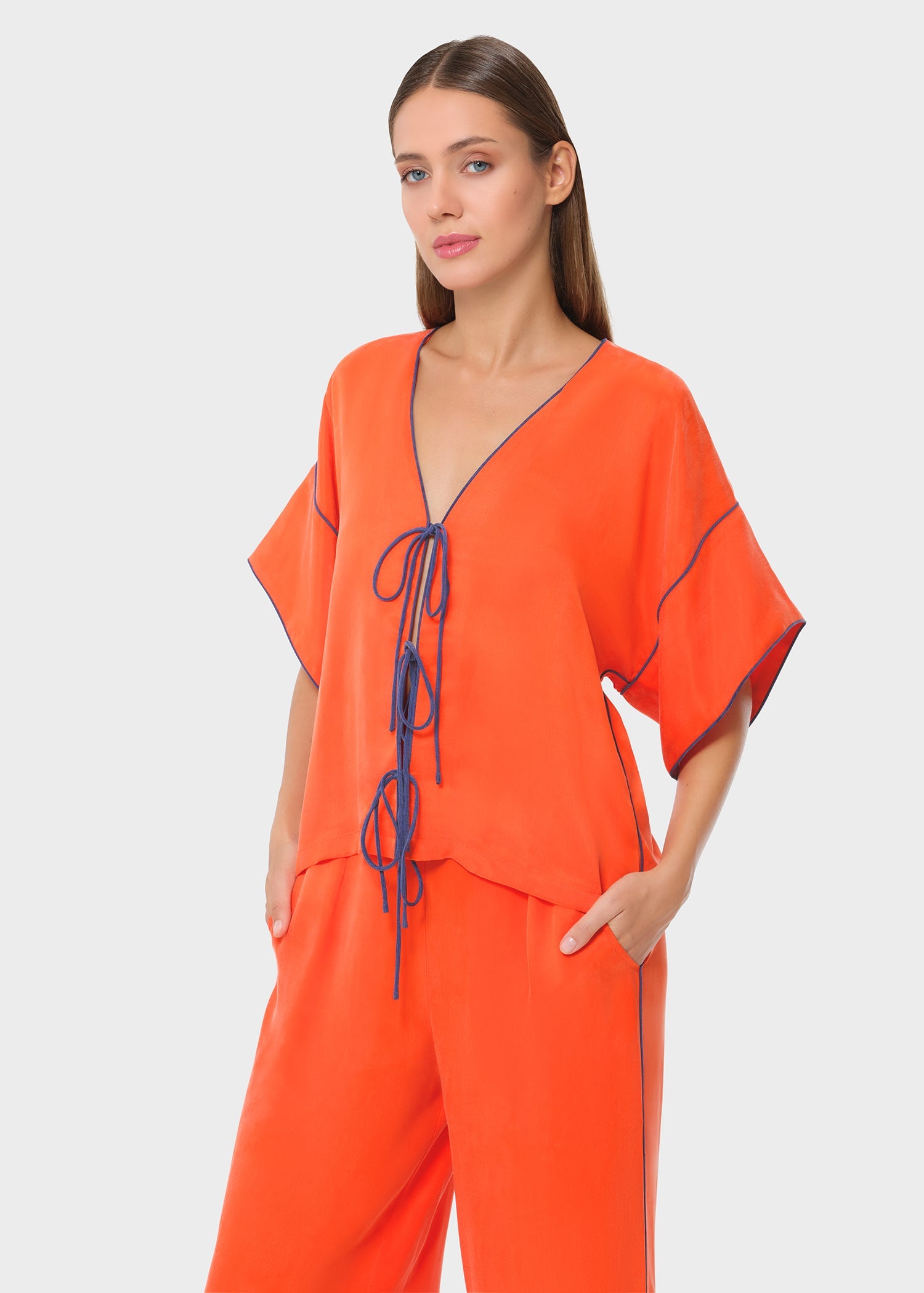 Close-up of a bright orange V-neck top with contrast piping and tie-up detail, paired with matching relaxed-fit shorts. Lightweight, flowy silhouette for an effortless look.