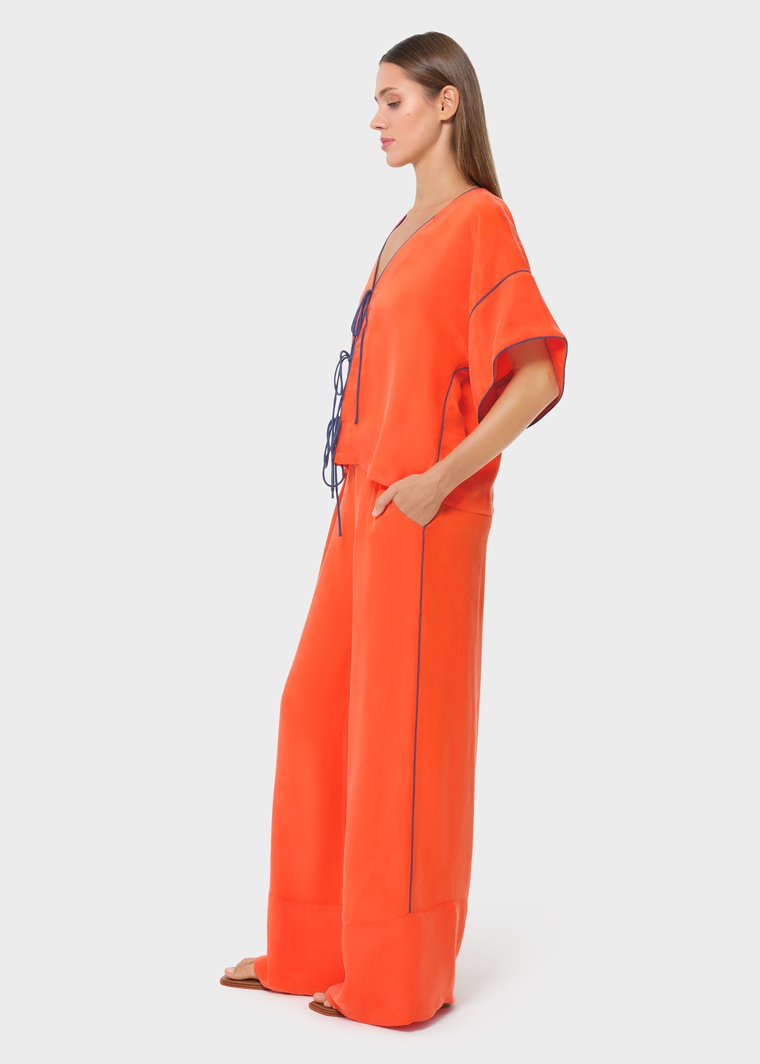Side view of a bright orange co-ord set with a relaxed V-neck top, contrast piping and tie-up detail, paired with wide-leg flowy pants for an effortless silhouette.