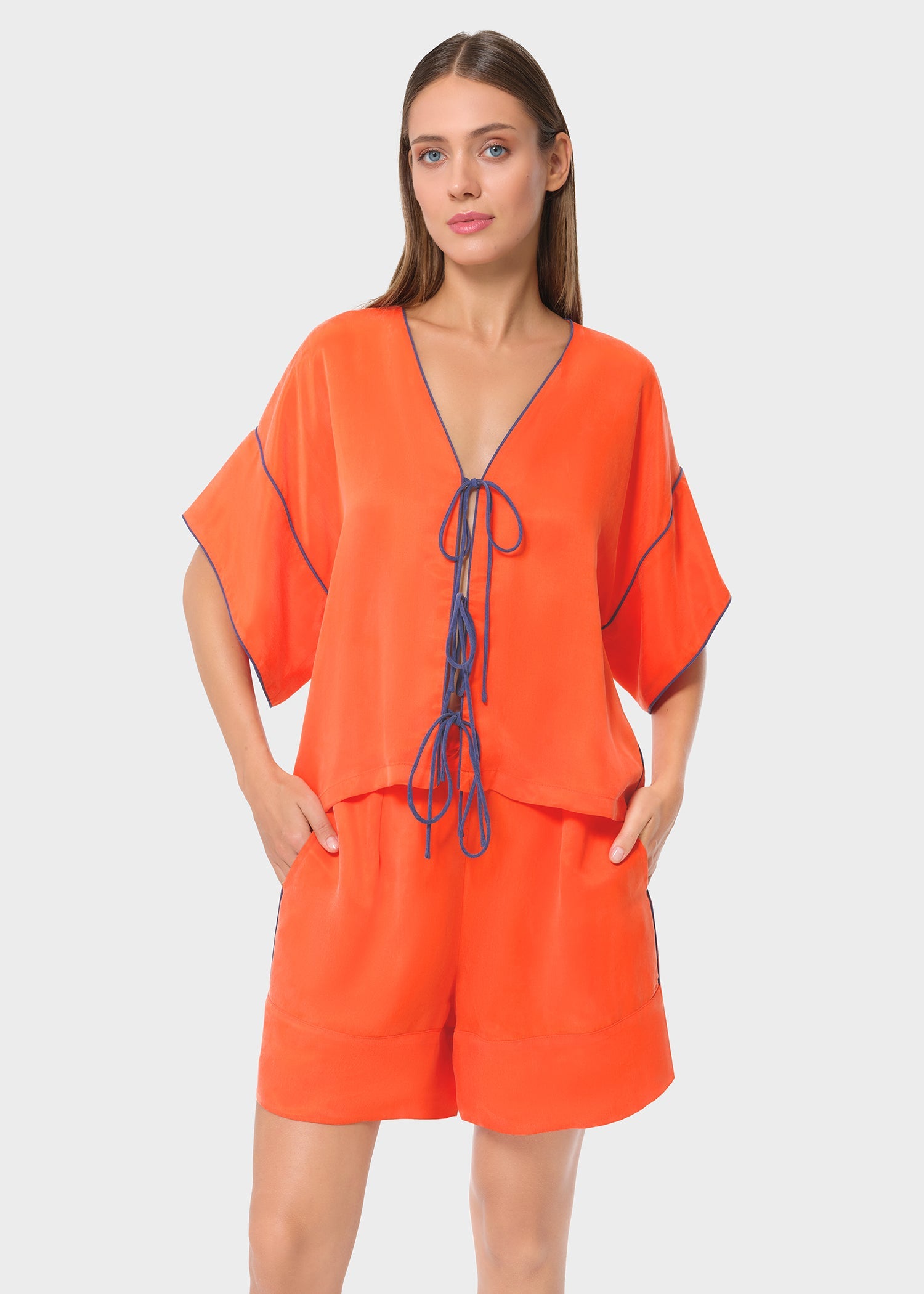 Front view of a bright orange co-ord set with a loose V-neck top featuring contrast piping and tie-up detail. Paired with matching relaxed-fit shorts for an easy, breezy look.