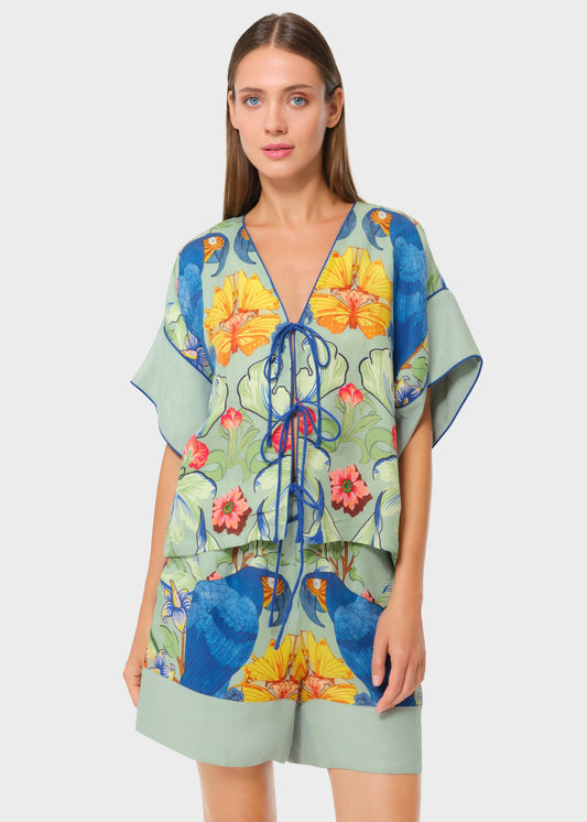 Model wearing a tropical print tie-front top and matching shorts featuring vibrant blue parrots and floral motifs on a soft green base, styled against a neutral studio background.