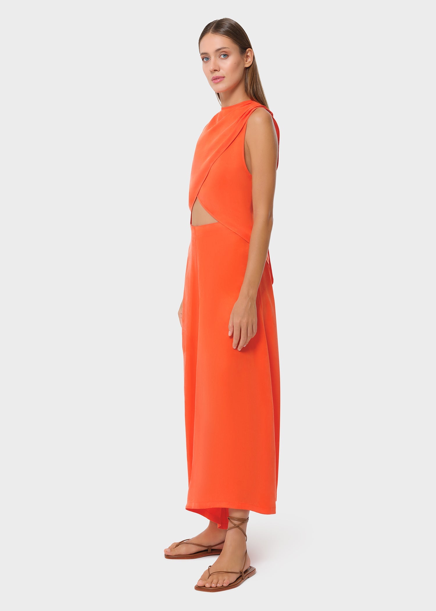 Side view of the vibrant orange sleeveless wrap top with soft draping and a subtle waist cutout. Paired with flowing wide-leg pants and flat sandals for a sleek, statement look.