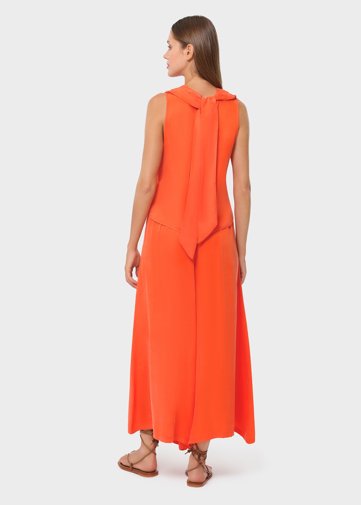 Back view of the vibrant orange sleeveless wrap top featuring a soft tie detail at the neckline. Paired with flowing wide-leg pants and flat sandals for an elegant, modern finish.