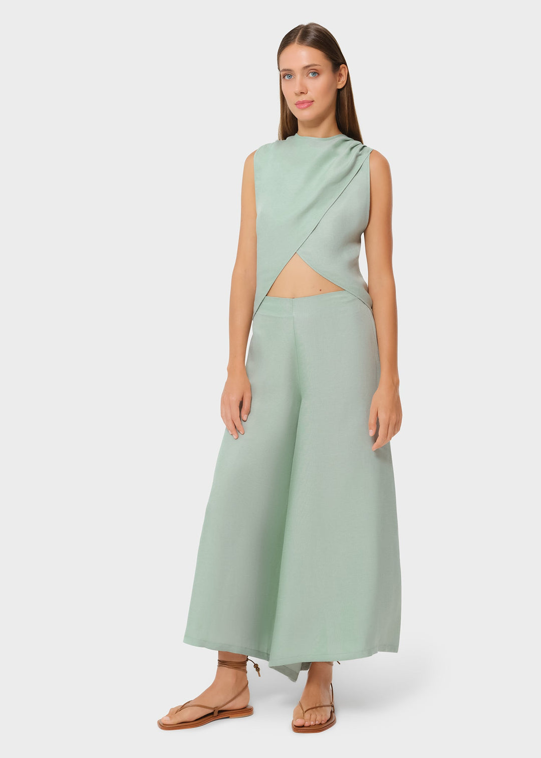 Woman wearing a mint green sleeveless asymmetrical wrap top with a subtle midriff cutout, paired with matching wide-leg pants and flat sandals, styled in a minimalist studio setting.
