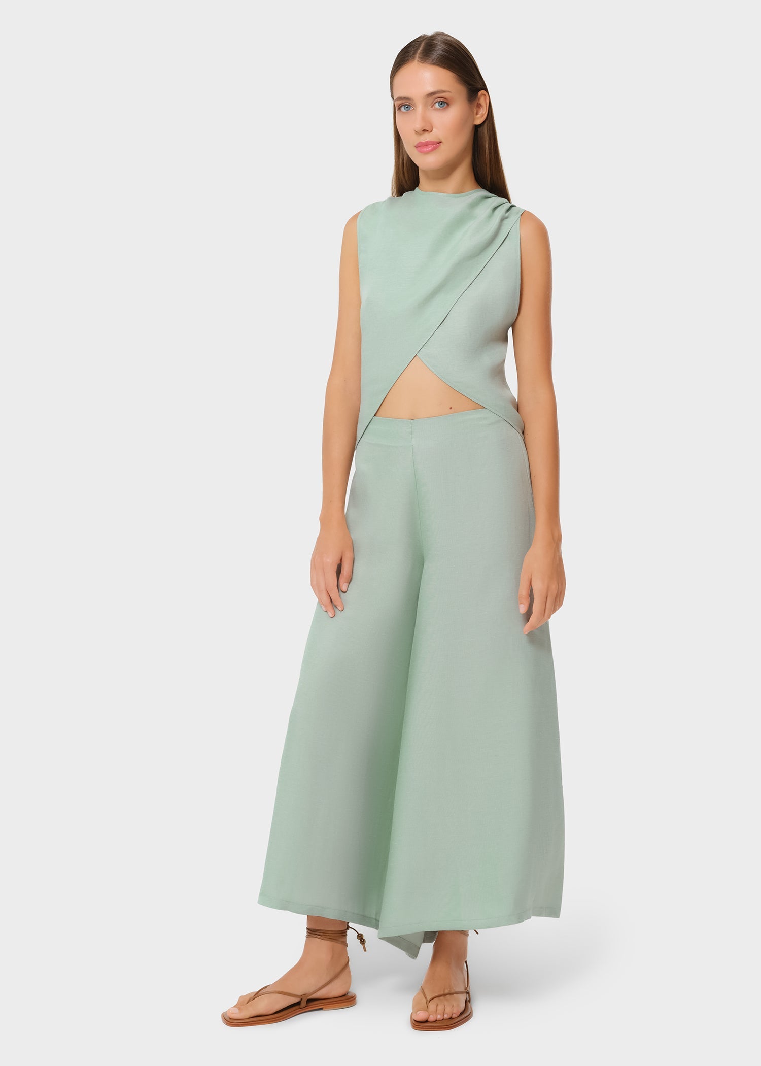Woman wearing a mint green sleeveless asymmetrical wrap top with a subtle midriff cutout, paired with matching wide-leg pants and flat sandals, styled in a minimalist studio setting.
