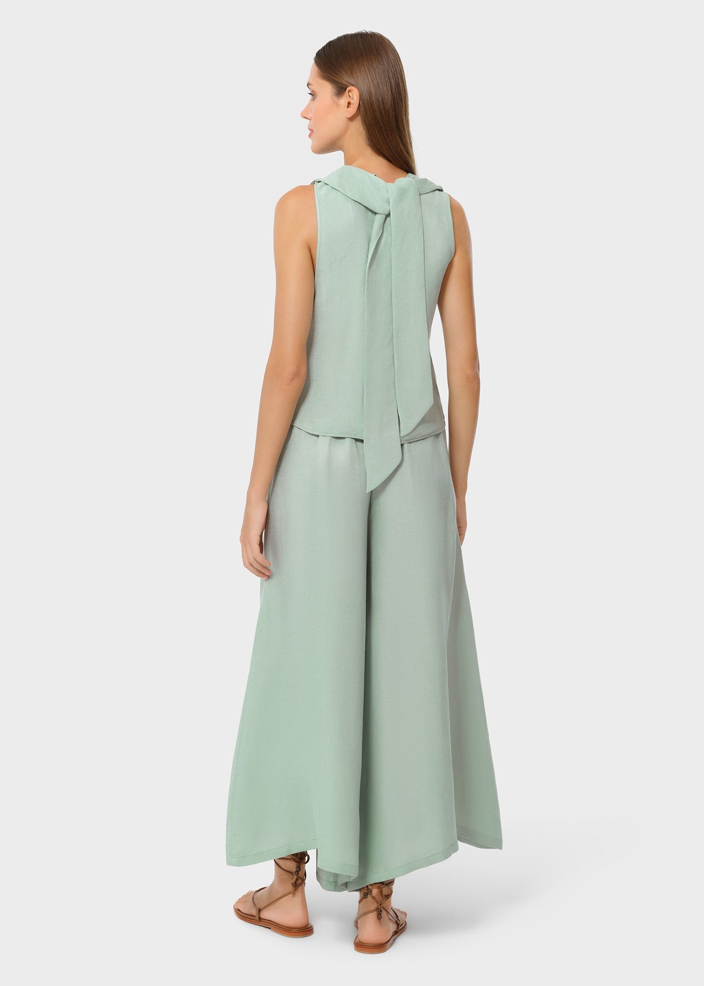 Back view of a woman in a mint sleeveless top with a tie detail at the neckline and matching wide-leg pants, styled with flat sandals in a studio setting.